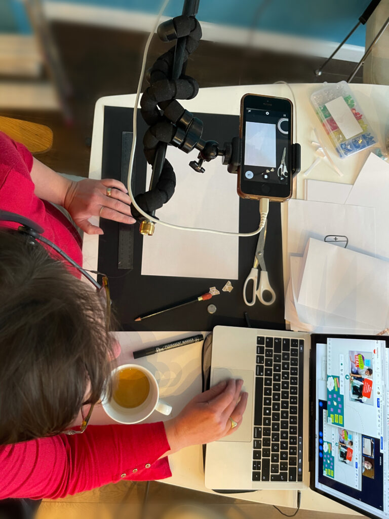 Digitale Workshop-Situation, von oben fotografierter Laptop, Handykamera und Person sowie Bastelmaterial